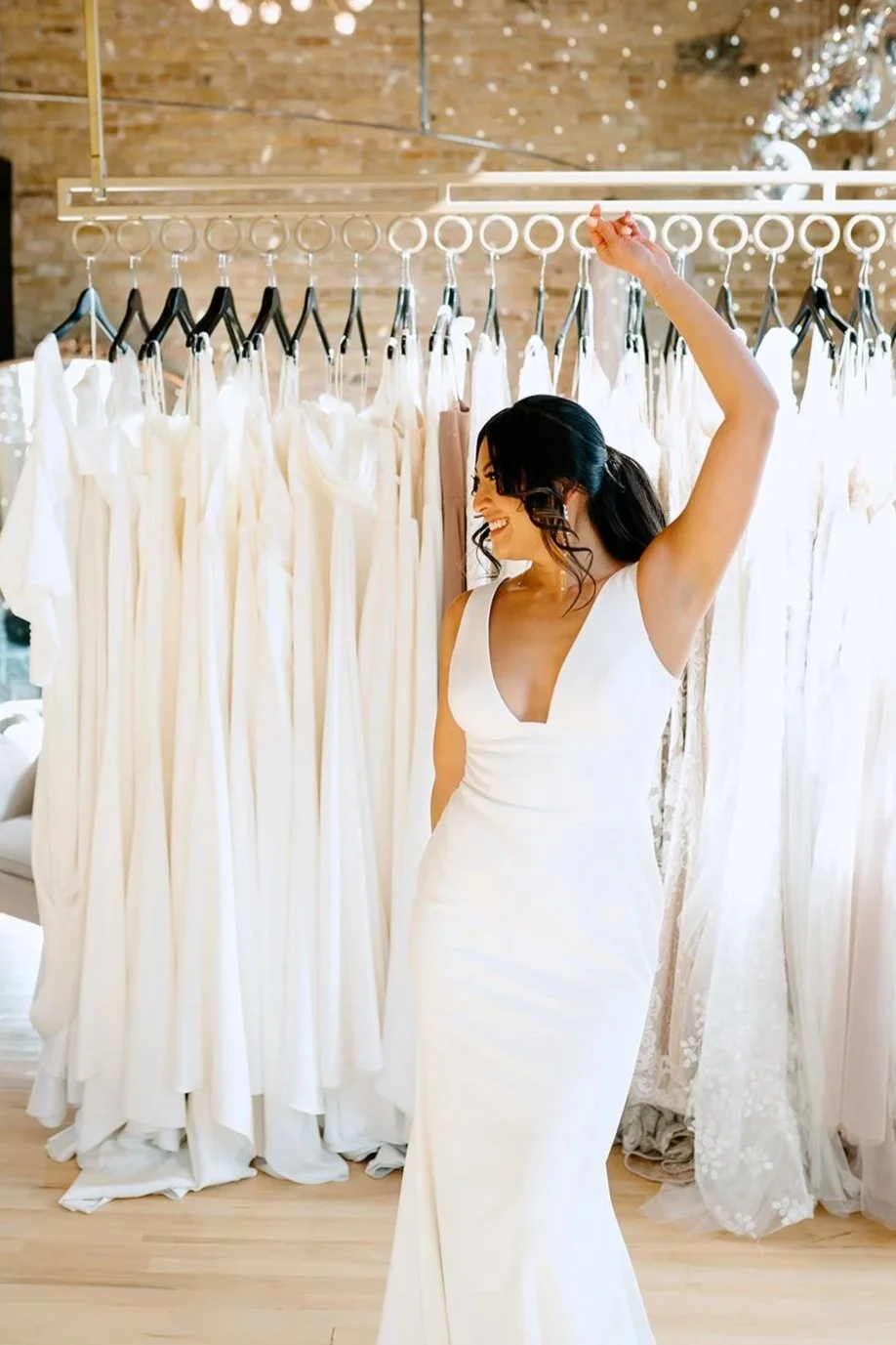 A woman in a white wedding dress is smiling and raising her arm in a bridal boutique with a rack of white wedding gowns behind her.
