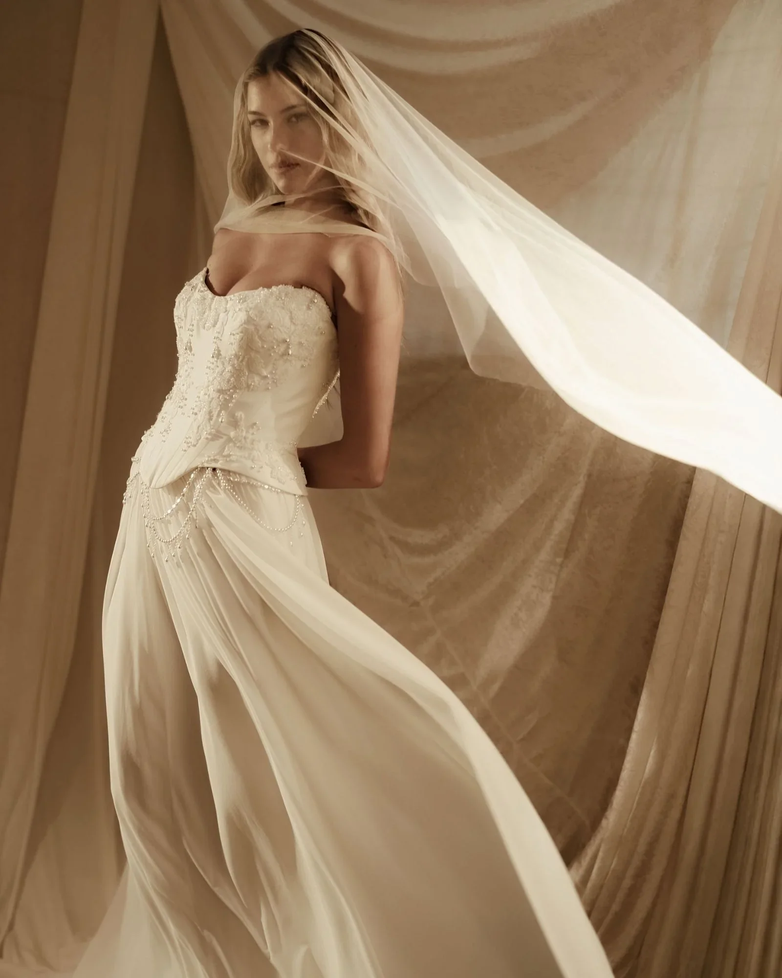 A woman in a white wedding dress with lace details, partially covered by a veil, standing in front of a mirror with light rays shining through.