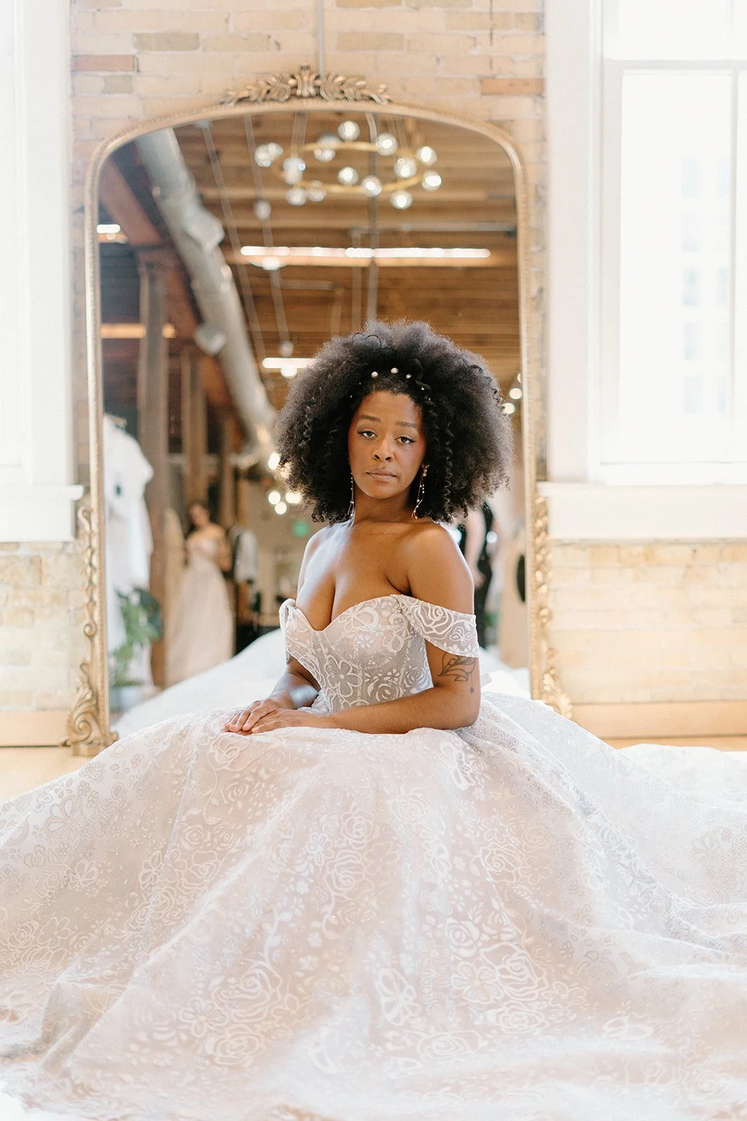 A woman in an off-shoulder wedding dress sitting in front of a mirror in a rustic room, with people and wedding gowns visible in the background.