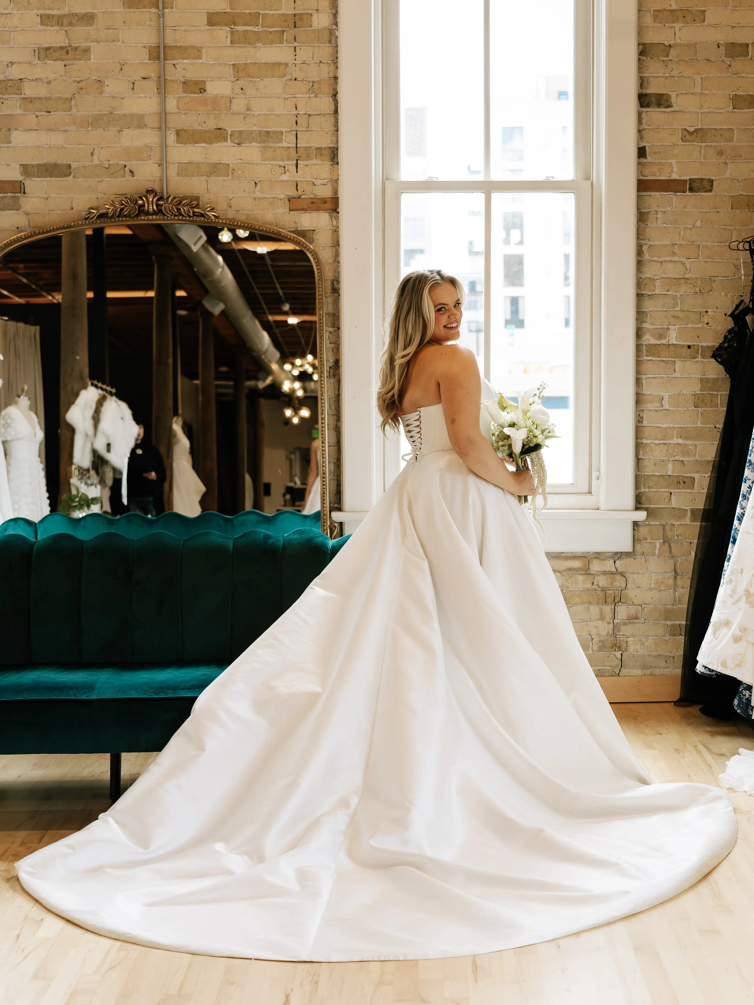 A smiling woman in an elegant wedding gown holding a bouquet of white flowers inside a boutique with brick walls and a large window.