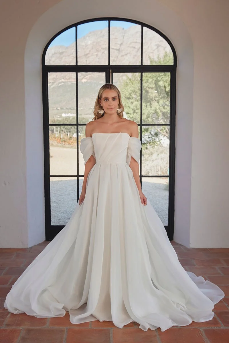 A woman in a white off-the-shoulder wedding gown standing in front of a large arched window with a scenic mountain and landscape view.