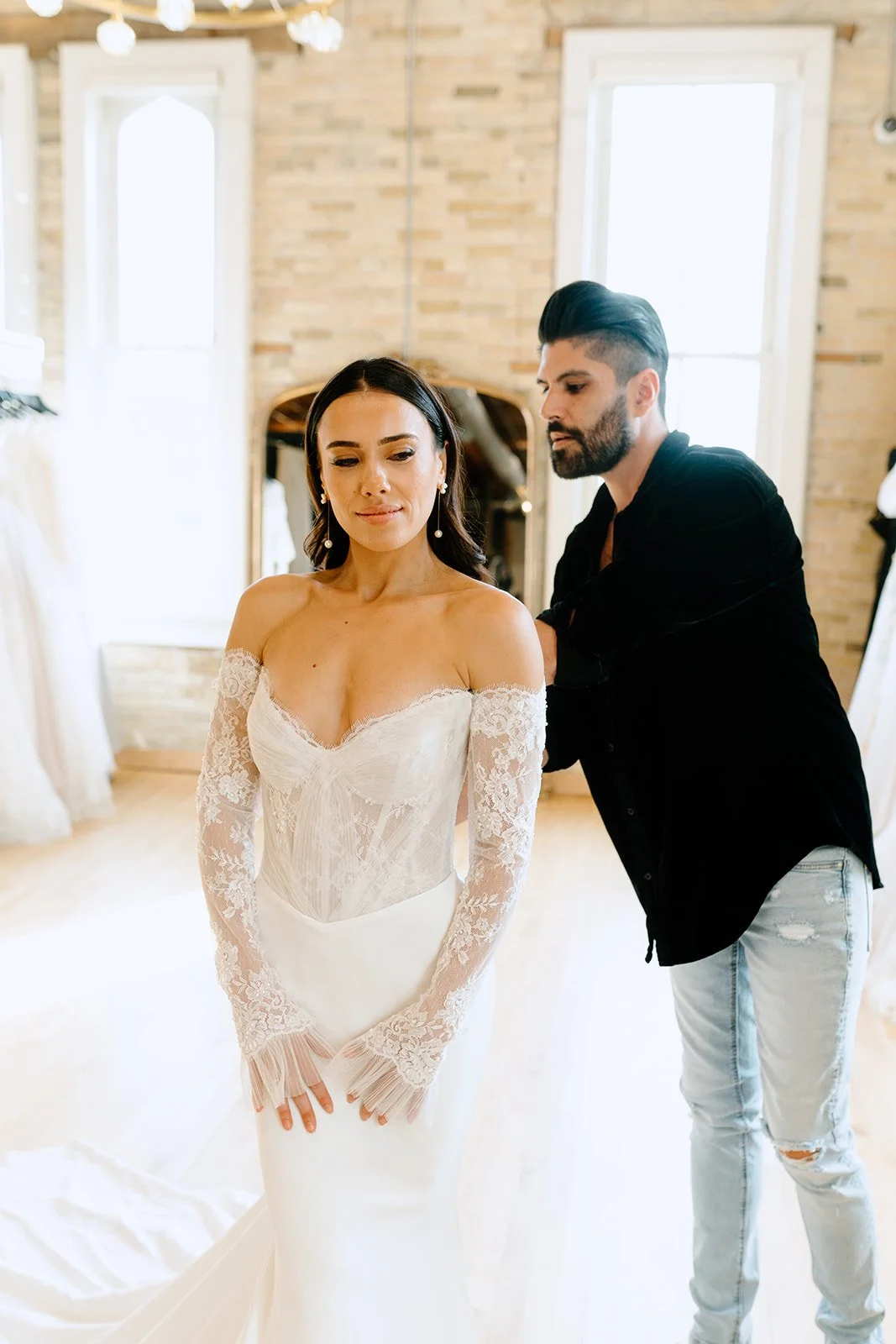 A woman in a wedding dress being assisted by a man in a black shirt, in a bright room with large windows and a mirror.