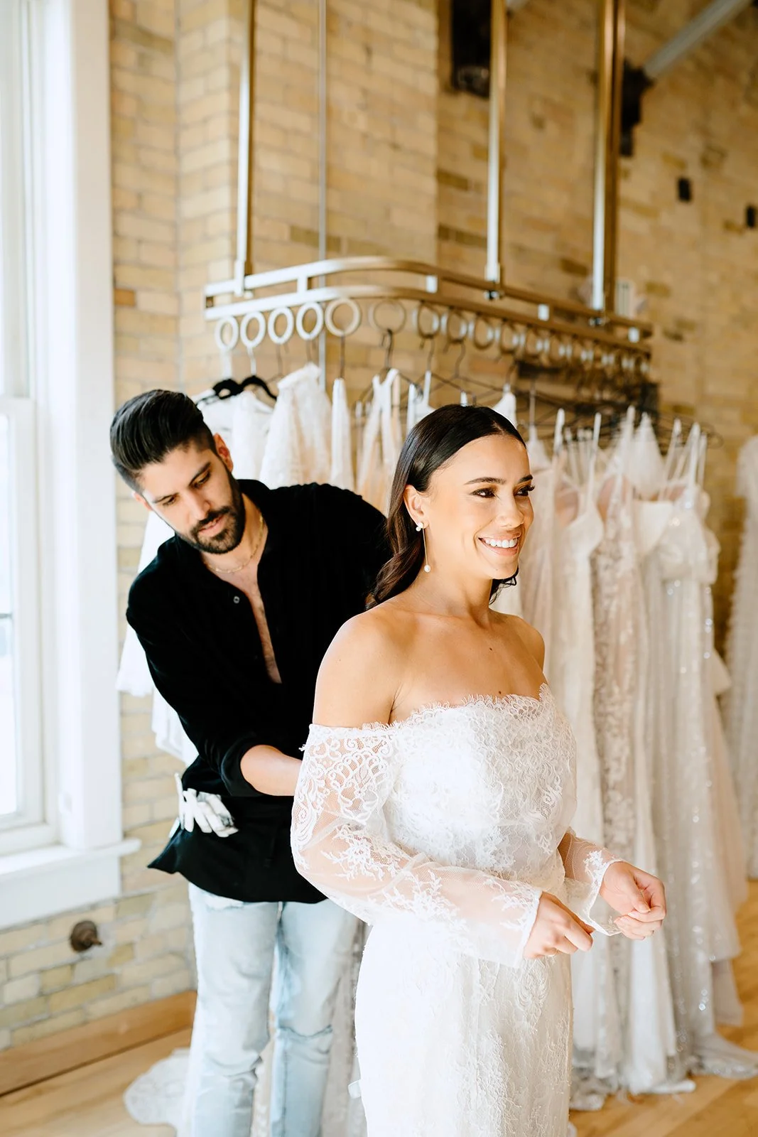 A woman in a white lace wedding dress smiling as she gets ready with a man helping her, surrounded by hanging wedding dresses in a room with brick walls.