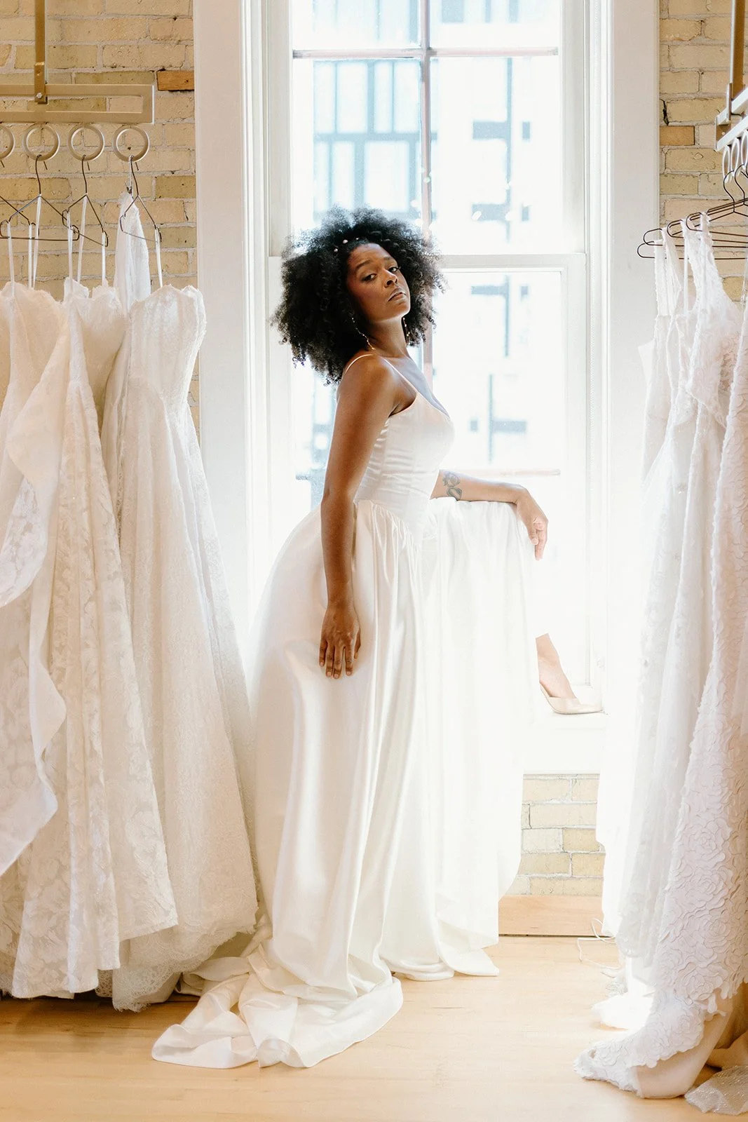 A woman in a white dress standing among rows of white wedding gowns in a bridal boutique, natural sunlight coming through large window.