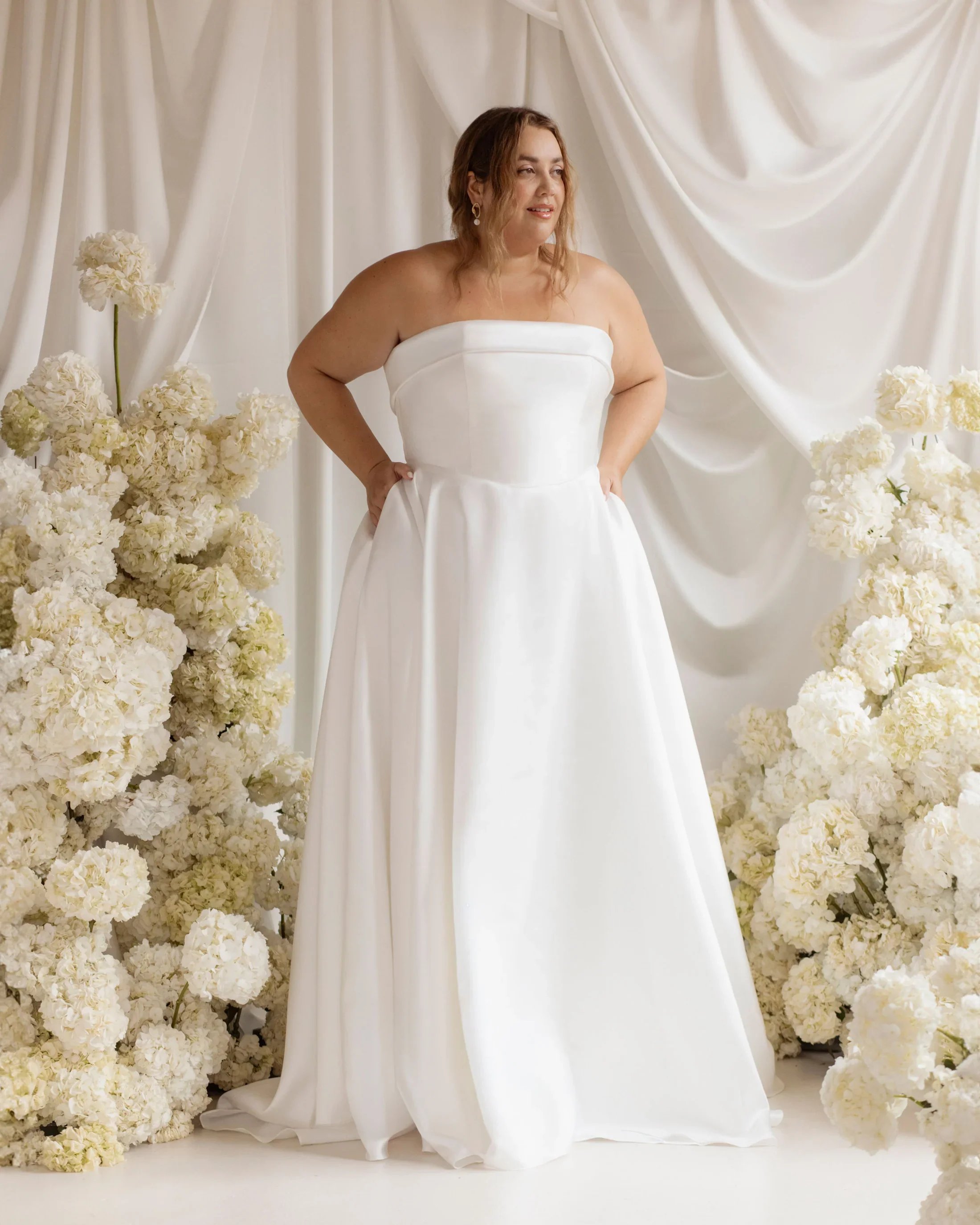 Woman in a white strapless wedding dress standing in front of a backdrop of white draped fabric and surrounded by white hydrangea flowers.