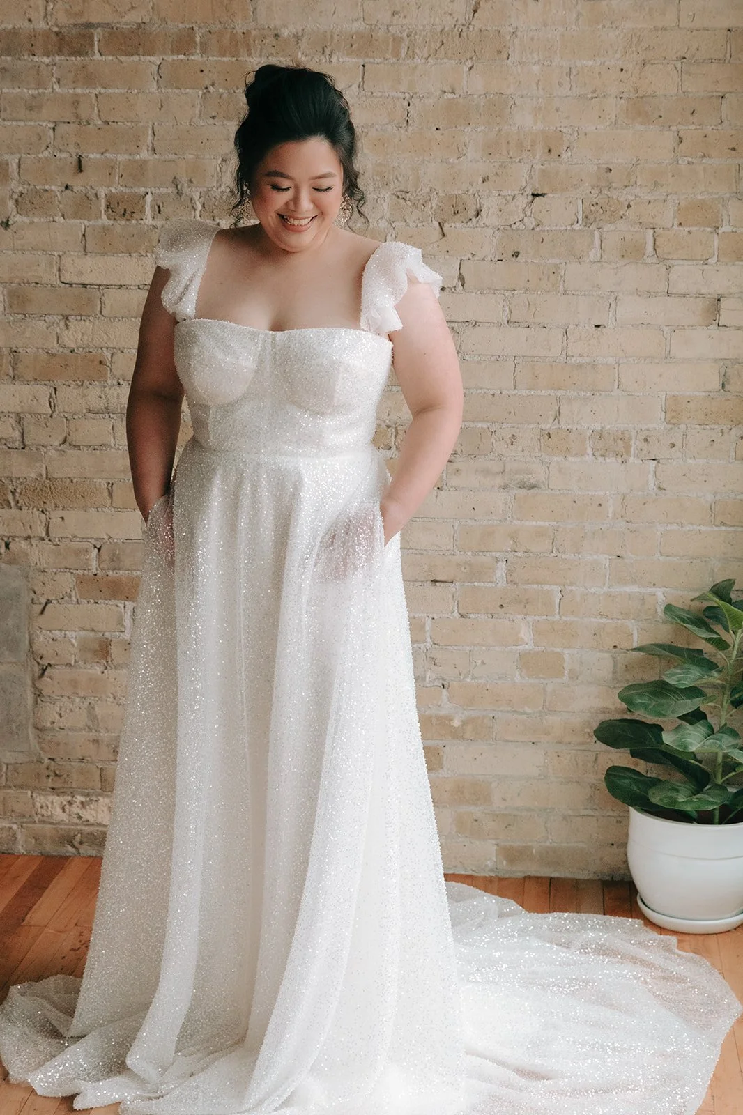 Woman in a white, sparkly gown standing in front of a brick wall with a potted plant nearby, smiling.