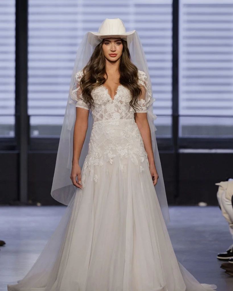 Woman wearing a wedding dress with a lace bodice and a flowy skirt, accessorized with a white hat and veil, standing indoors.