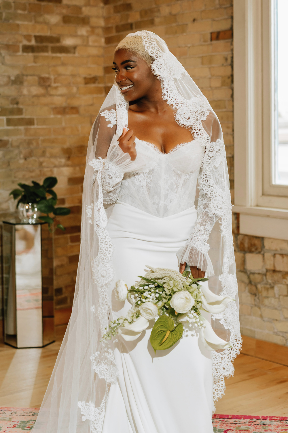 A bride with short platinum blonde hair wearing a white lace wedding dress and veil, holding a floral bouquet, smiling indoors with brick wall background and window.