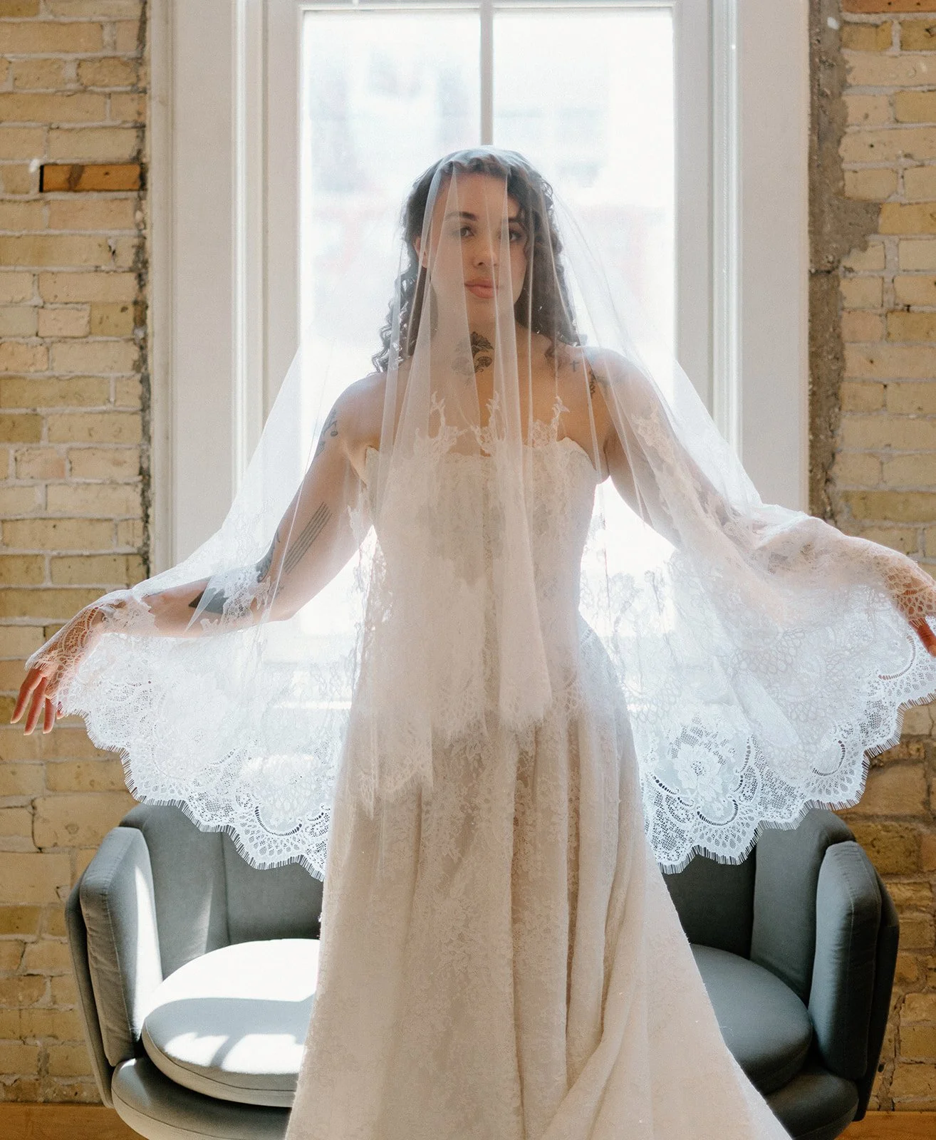 A woman in a lace wedding dress and veil standing in front of a large window, with a brick wall behind her.
