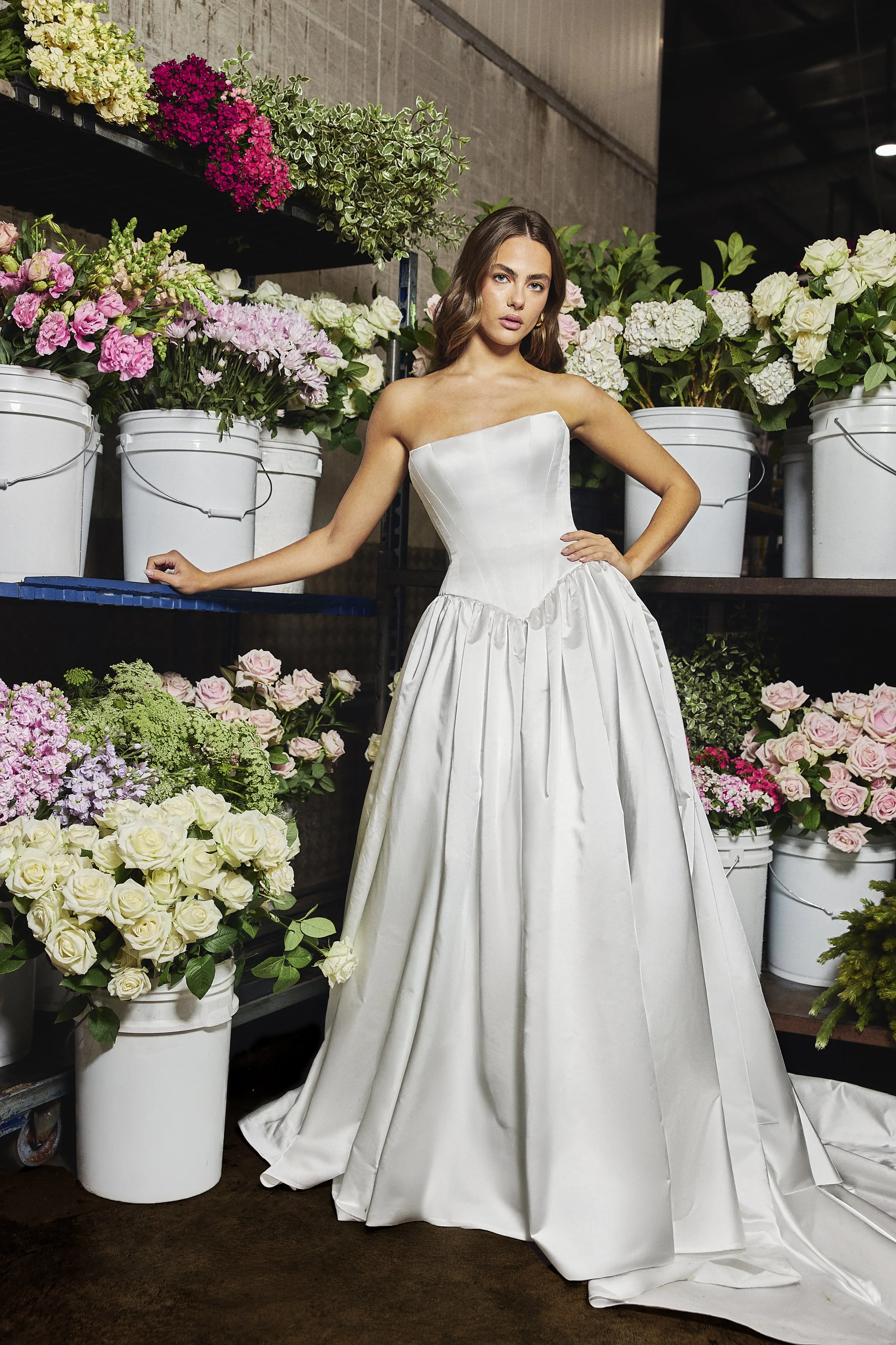 A woman in a strapless white wedding gown standing in a flower shop surrounded by buckets of pink, white, and purple flowers.