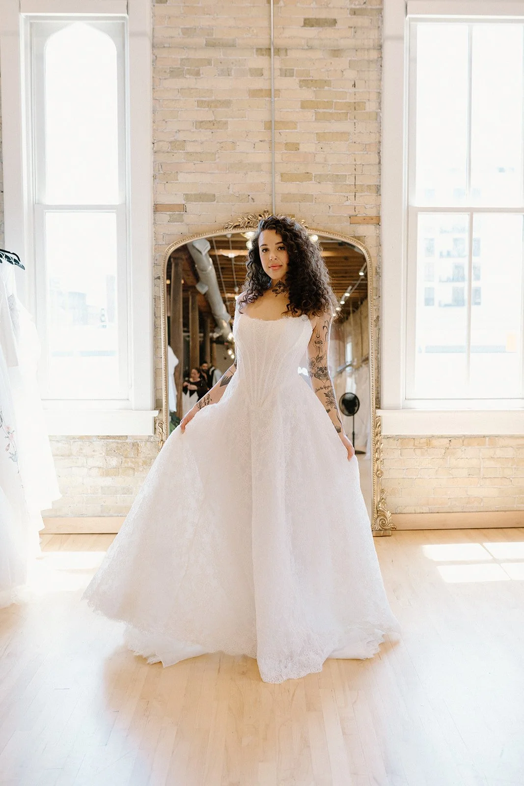 A woman with curly hair and tattoos on her arms is wearing a white wedding dress in front of a large mirror with a gold frame in a bright room with brick walls and large windows.