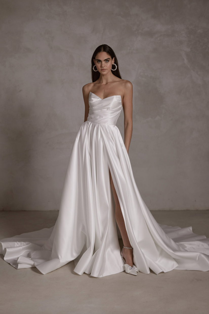 A woman wearing a strapless white satin wedding dress with a high slit, standing against a plain gray wall.
