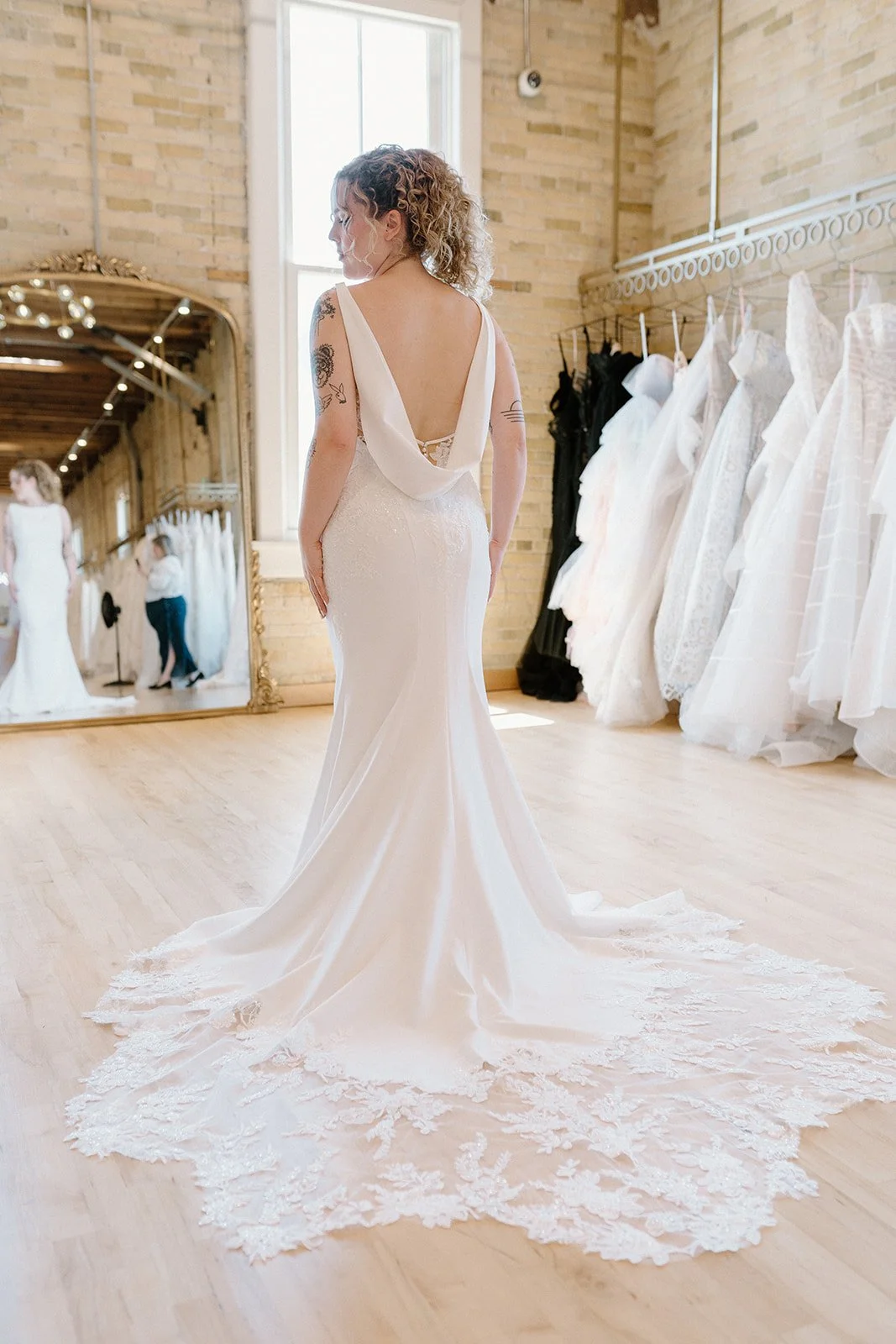 A woman in a wedding dress trying on the gown in a bridal shop, reflected in a large mirror, with extra wedding dresses on display and a bright window in the background.