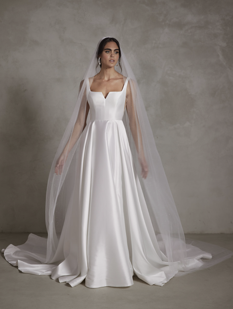 Bride in a white wedding gown with a fitted bodice, square neckline, and full satin skirt, wearing a long veil and earrings, standing against a plain gray background.