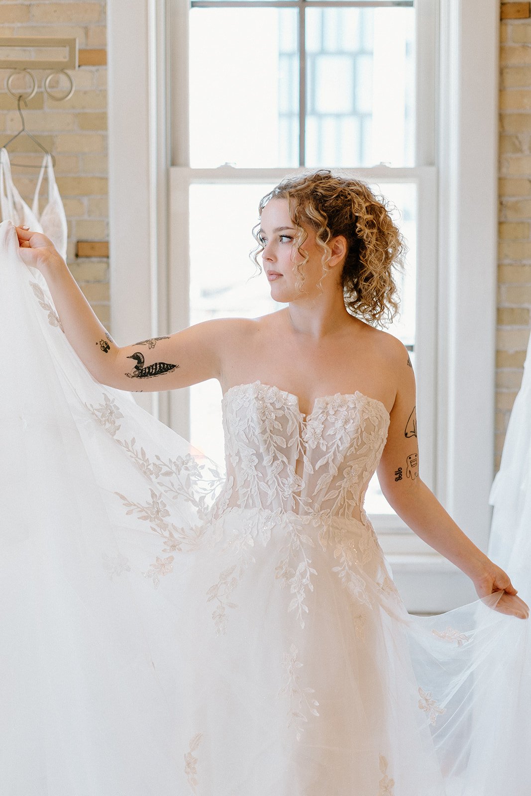 A woman with curly hair and tattoos wearing a strapless wedding dress, holding the fabric of her dress near a window.