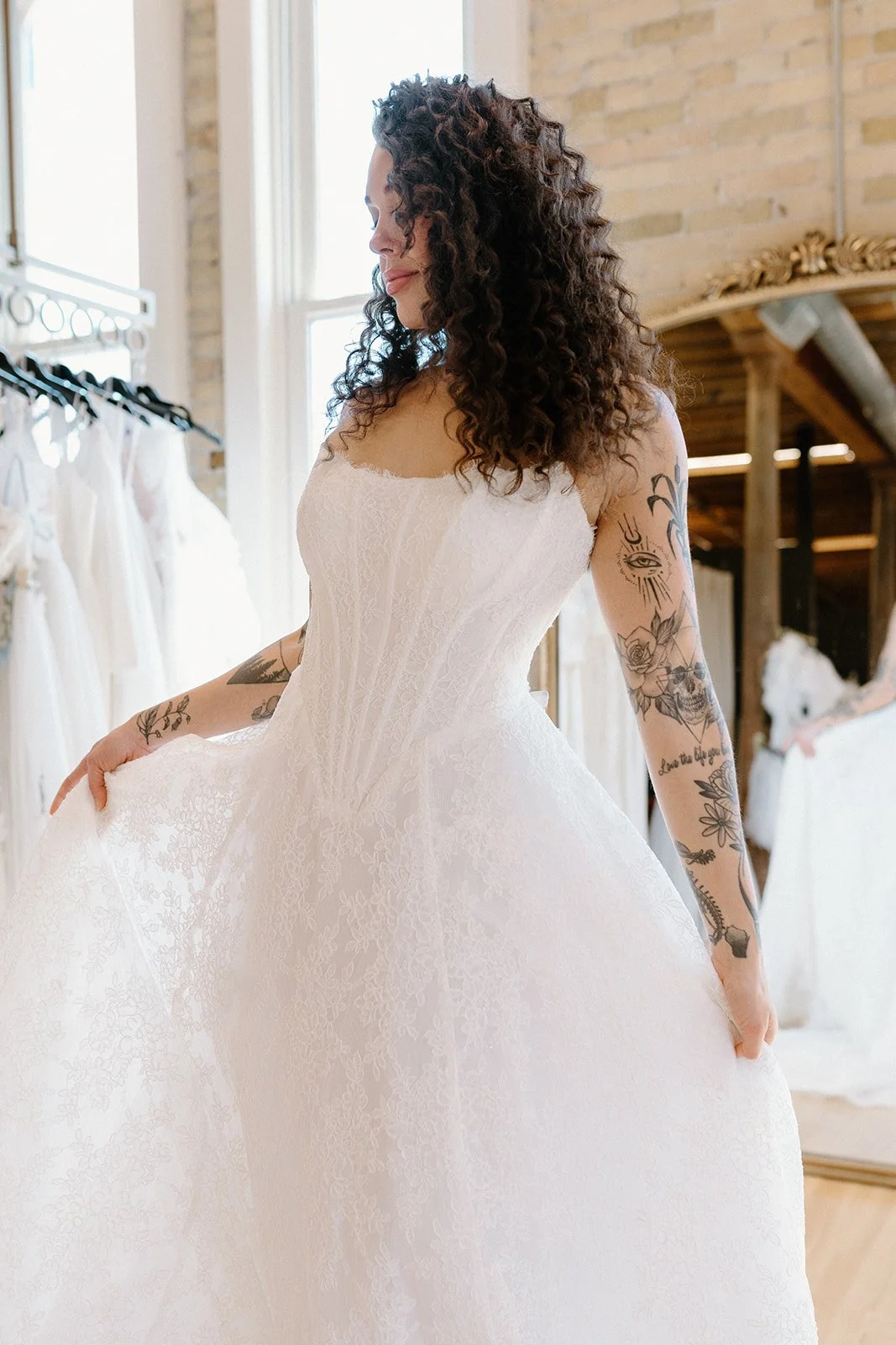 A woman with curly dark hair, wearing a white lace dress, standing in a bright room with exposed brick wall. She has tattoos on her arms and is holding the skirt of her dress, smiling softly.