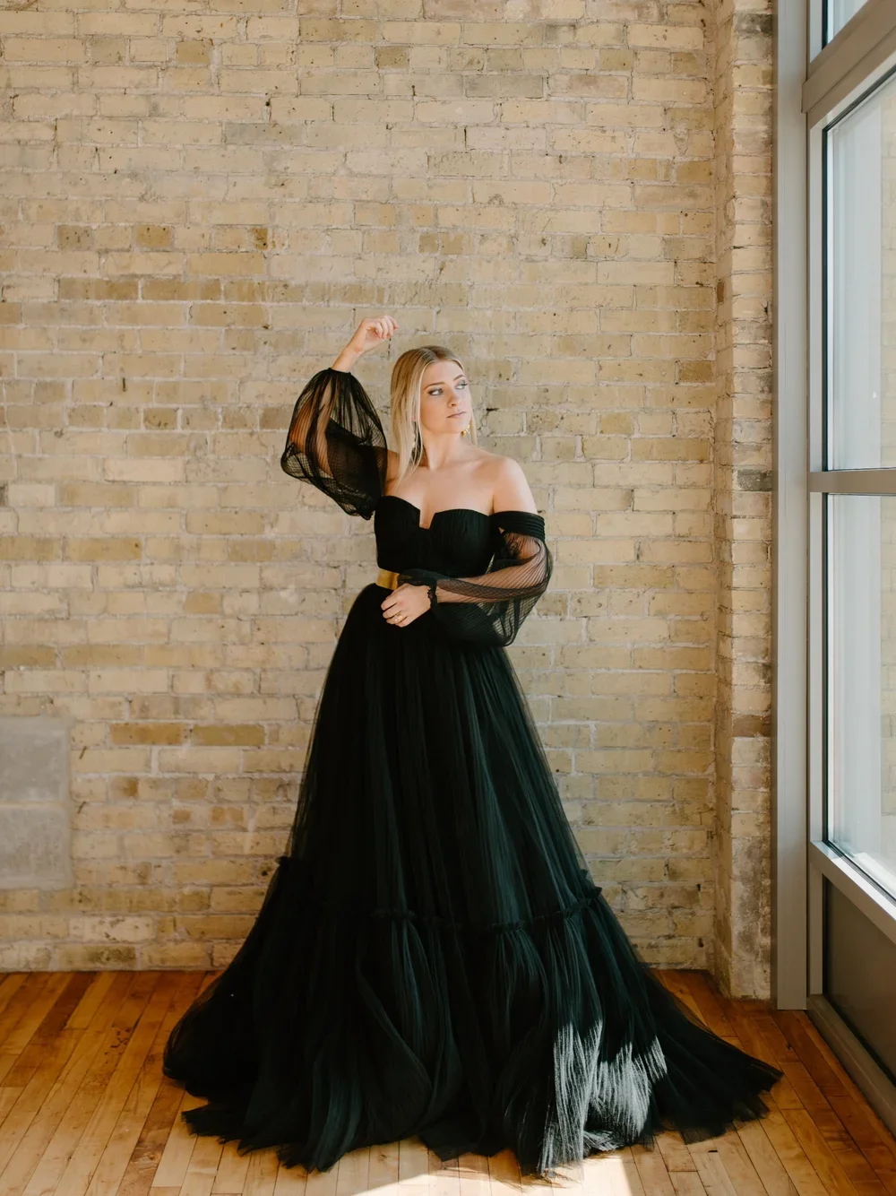 A woman in a long black gown with sheer off-the-shoulder sleeves stands beside a large window with a brick wall in the background.