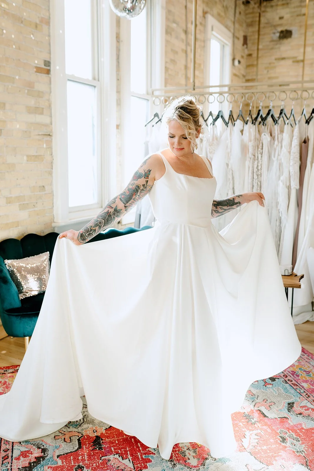 A woman in a white wedding dress holding out the sides of her skirt, indoors with large windows and a rack of wedding dresses behind her.
