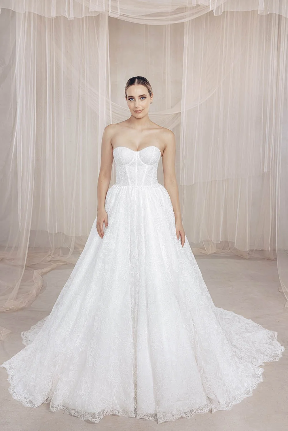 A woman wearing a strapless white wedding dress with lace details, standing in front of a backdrop with sheer fabric draped from the ceiling.