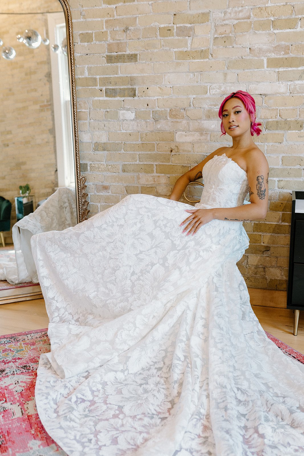 A woman with pink hair wearing a white lace strapless wedding dress, sitting on a chair against a brick wall, holding out the train of her dress.