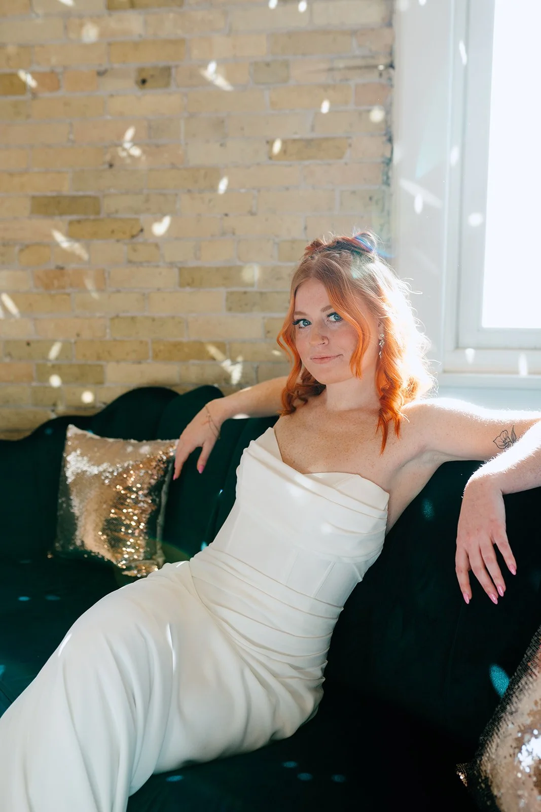 bride wearing a fitted crepe wedding dress with corset details leaning back with her arms on dark green couch with sparkle pillows next to a cream brick wall and window