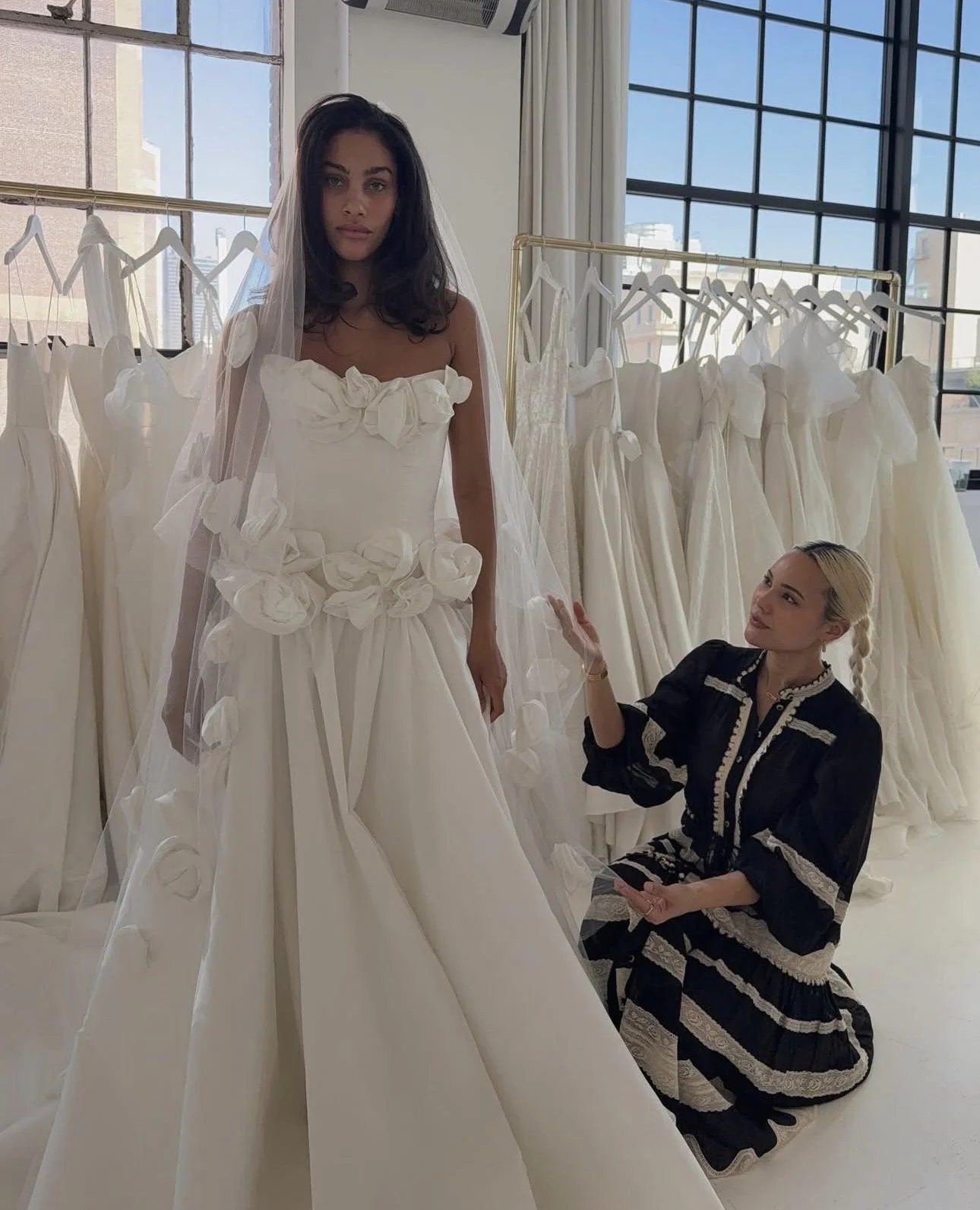 A woman trying on a wedding dress in a fitting room while another woman, designer, looks on and discusses the dress. The wedding dress is white with floral embellishments and a full skirt. In the background, there are racks of similar white wedding dresses and large windows letting in natural light.