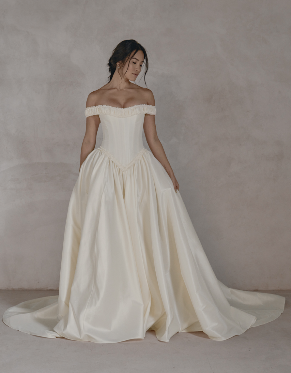 A woman in a strapless, off-the-shoulder, cream-colored wedding gown holding the skirt with one hand, standing against a light textured background.