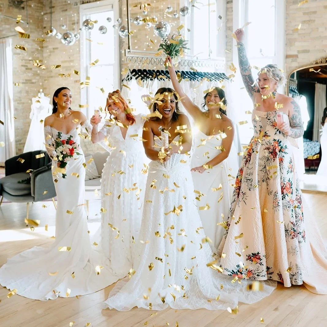 Group of women in wedding dresses celebrating with golden confetti in a bright room.