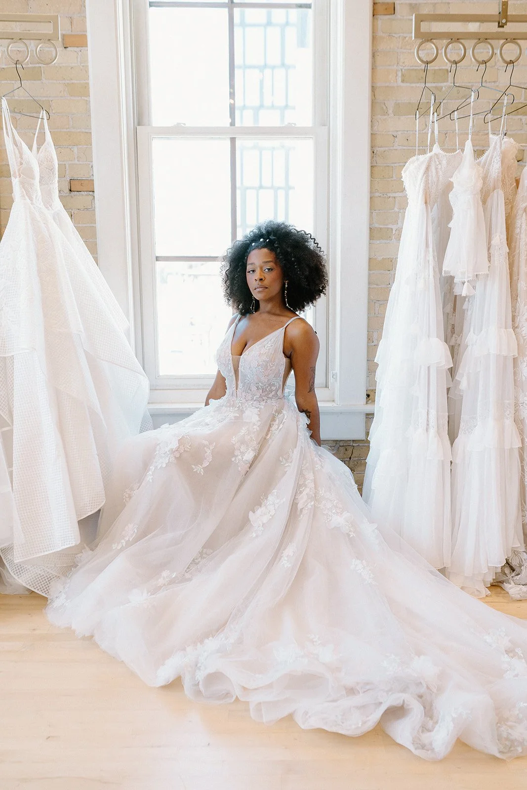 A woman in a wedding dress sitting in a bridal boutique with white dresses hanging on both sides.