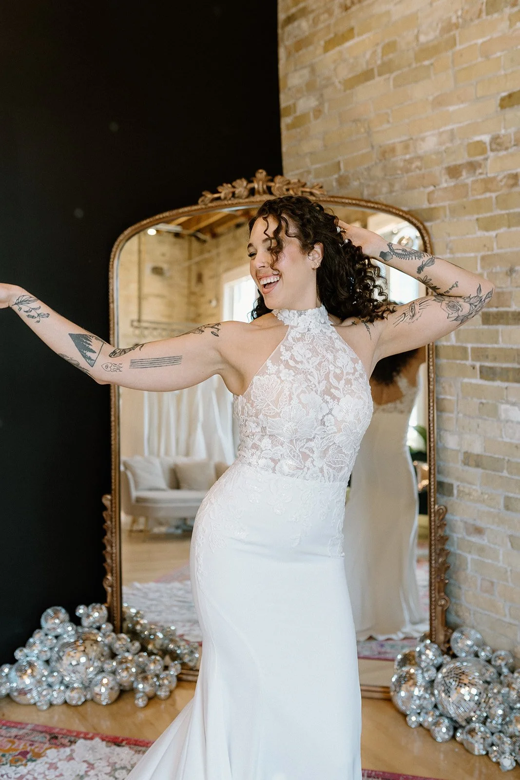 A woman wearing a white lace wedding dress, smiling and dancing in front of a large mirror, with decorative silver spheres on the floor.