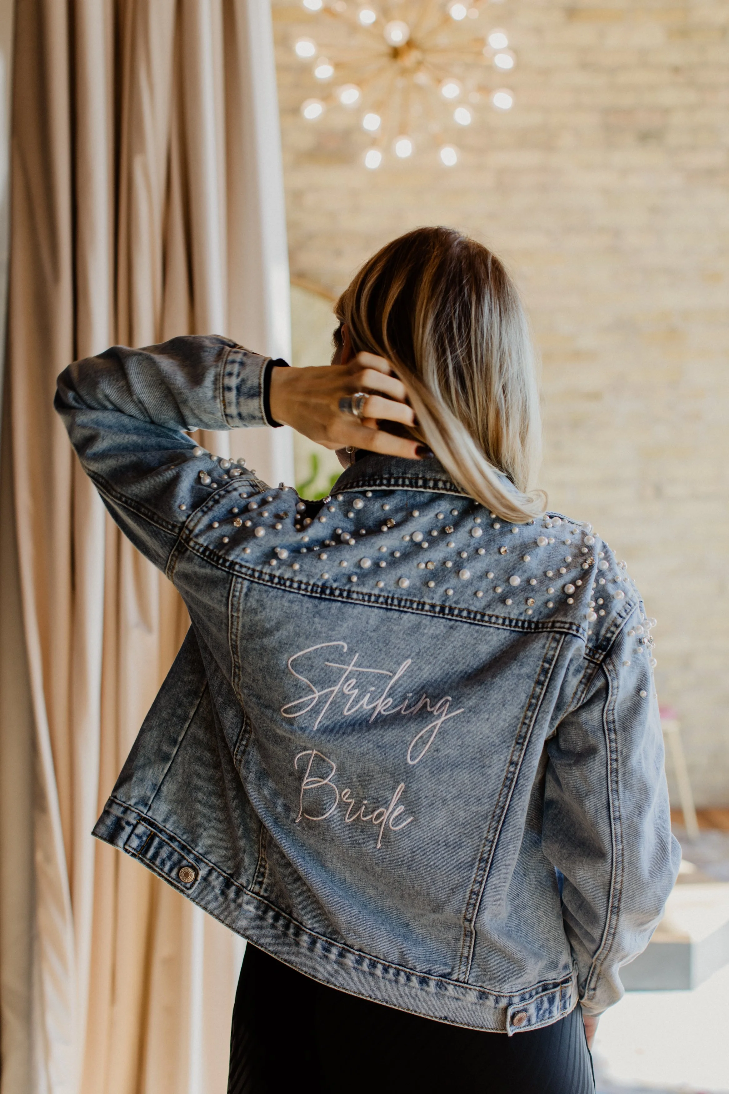 Back view of a woman in a denim jacket with pearl embellishments, featuring the embroidered phrase 'Striking Bride,' standing indoors near beige curtains and a brick wall with a chandelier.