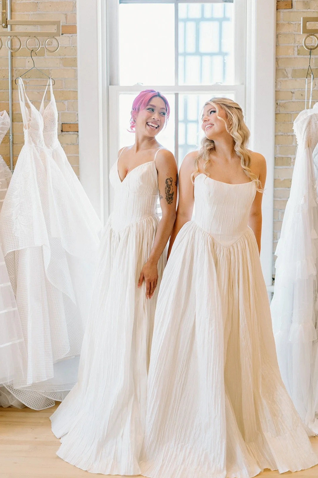 Two women in cream-colored dresses inside a bridal boutique, smiling and looking at each other, surrounded by hanging wedding dresses.