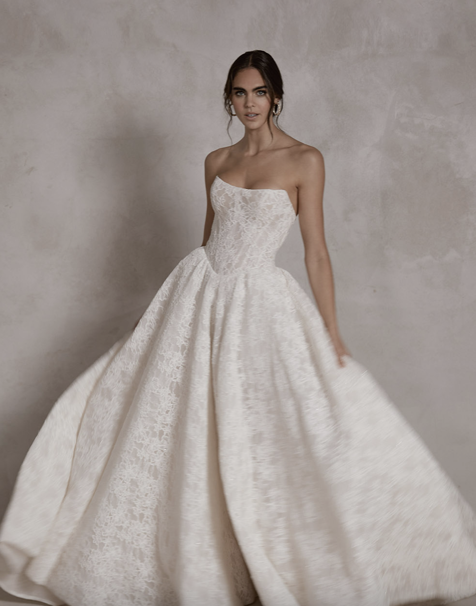 A woman in a strapless, white lace wedding gown with a voluminous skirt, standing against a neutral textured background.