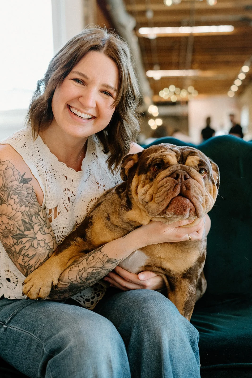 A woman with tattoos on her arm is smiling and holding a large bulldog. They are inside a well-lit room with a wooden ceiling, and there are blurred people in the background.