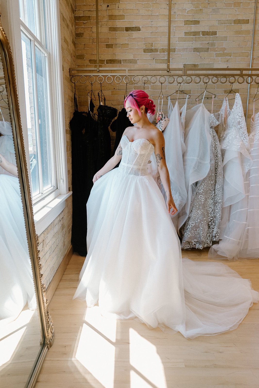 A woman with pink hair in a strapless wedding dress stands next to a mirror, looking down, in a bridal boutique with dresses hanging on a rack behind her.
