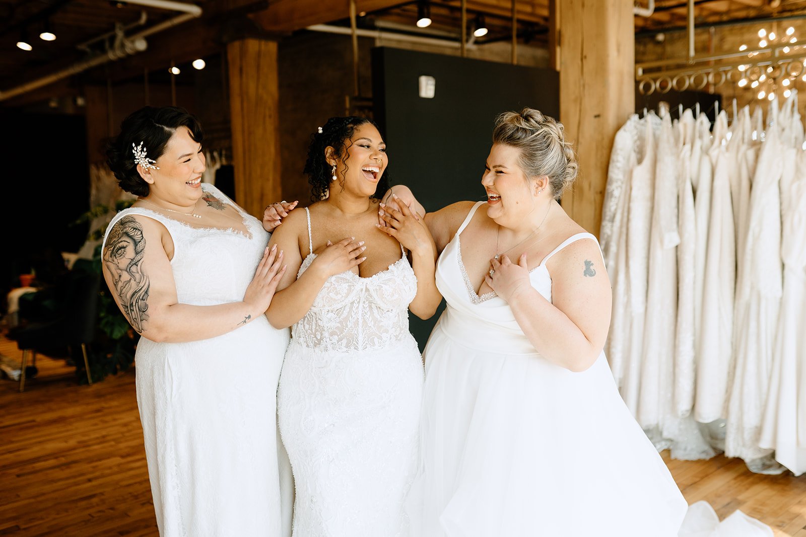 Three women in white wedding dresses sharing a joyful moment in a bridal shop, with wedding gowns on racks in the background.