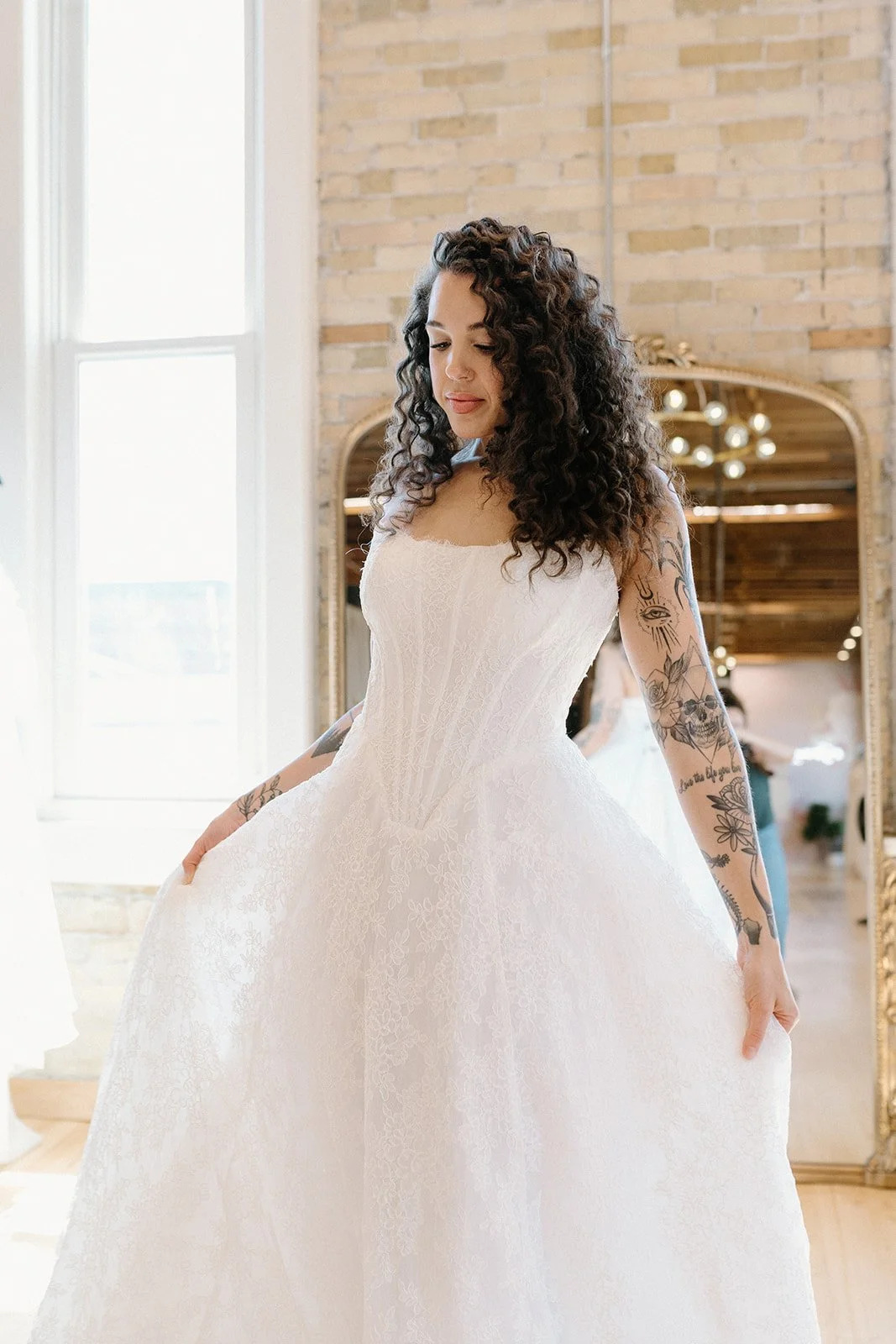 A young woman with curly dark hair wearing a white lace wedding dress, standing indoors near a window, looking down with a slight smile, with tattooed arms visible.