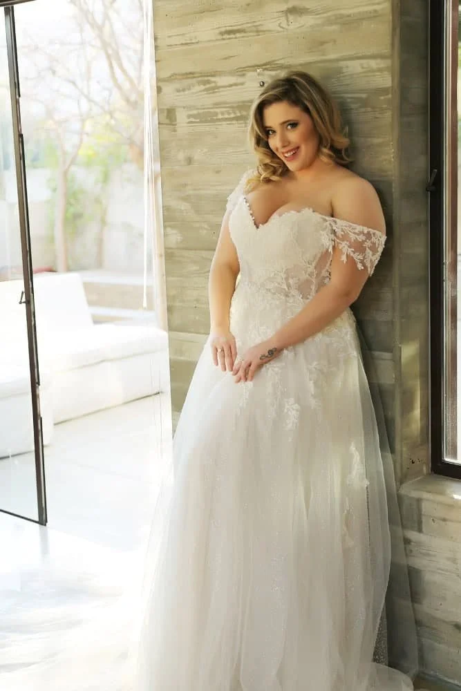 A woman in an off-the-shoulder white wedding dress with lace details, standing indoors near a window with a wooden wall and a white sofa in the background.