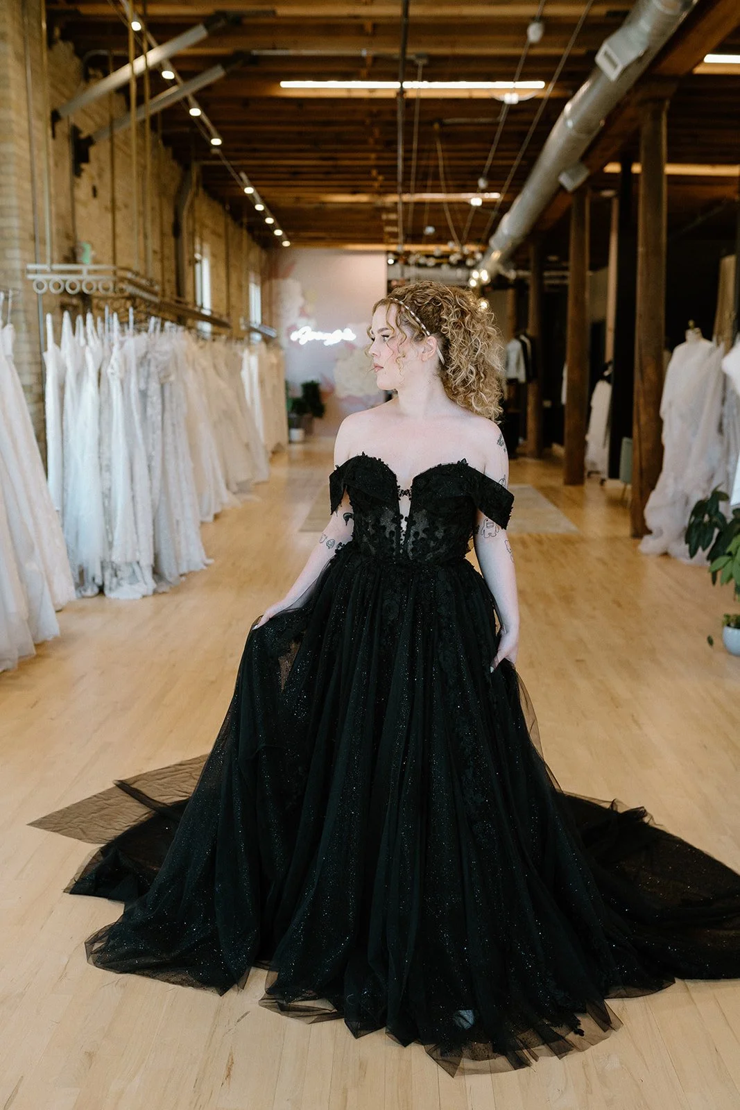 Woman trying on a black off-the-shoulder wedding gown inside a bridal boutique with dresses on racks and a neon sign in the background.
