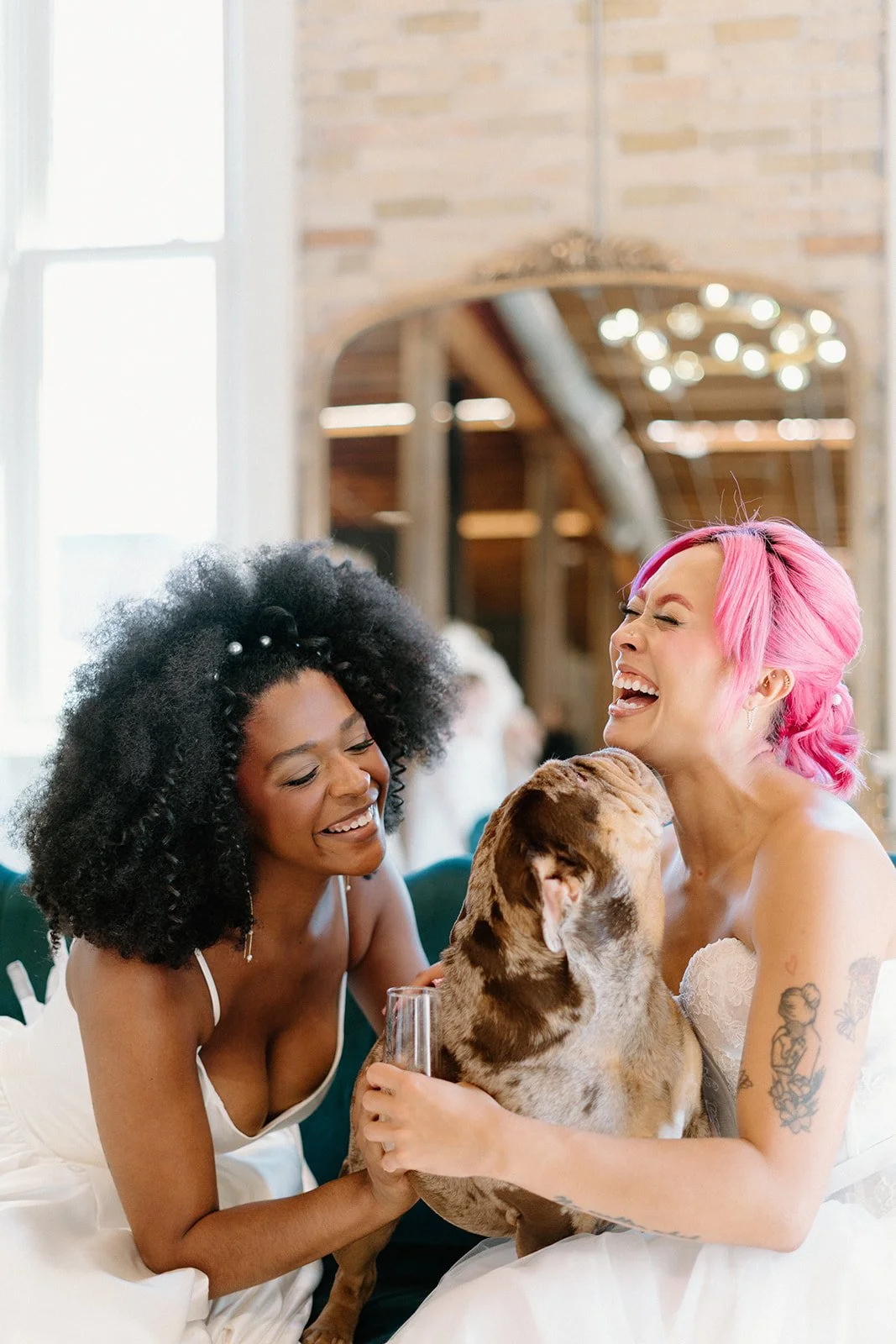 Two women laughing and holding a dog, sitting on a couch with a brick, indoor background.