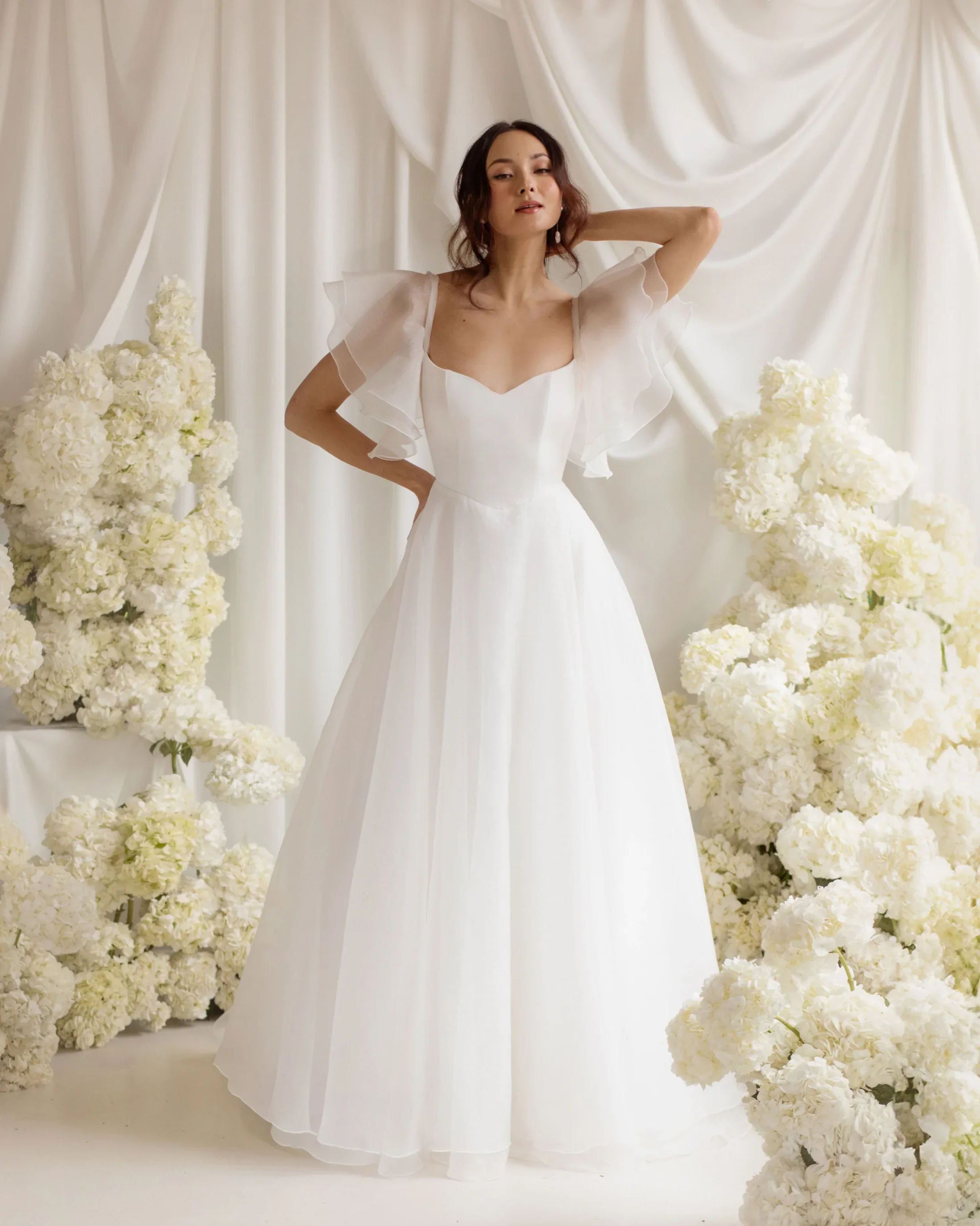 A woman in a white wedding dress standing between large white floral arrangements on a cream backdrop.