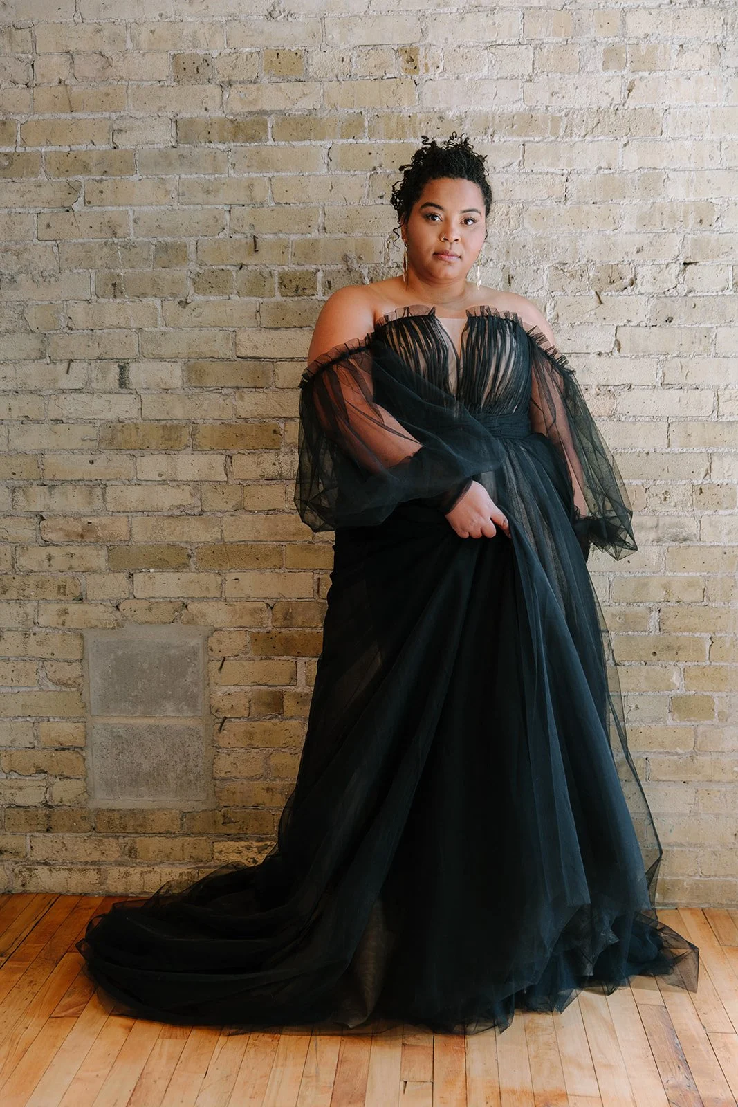 A woman in a black, off-shoulder evening gown with sheer, ruffled sleeves, standing against a beige brick wall.