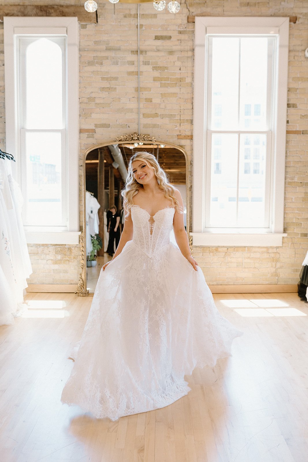 A woman in a wedding dress standing in front of a mirror in a bright room with brick walls and large windows.