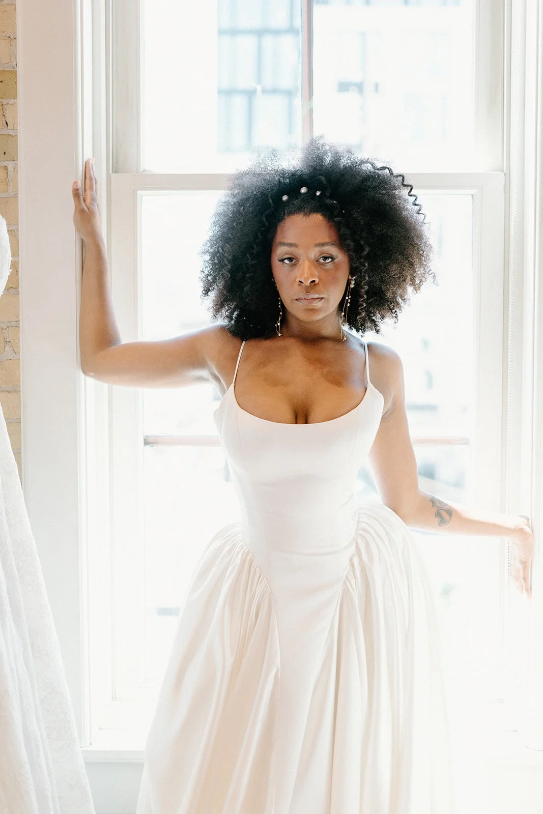 A woman with natural curly hair wearing a white dress, standing by a large window, with one arm resting on the window frame and the other lowered, looking directly at the camera.