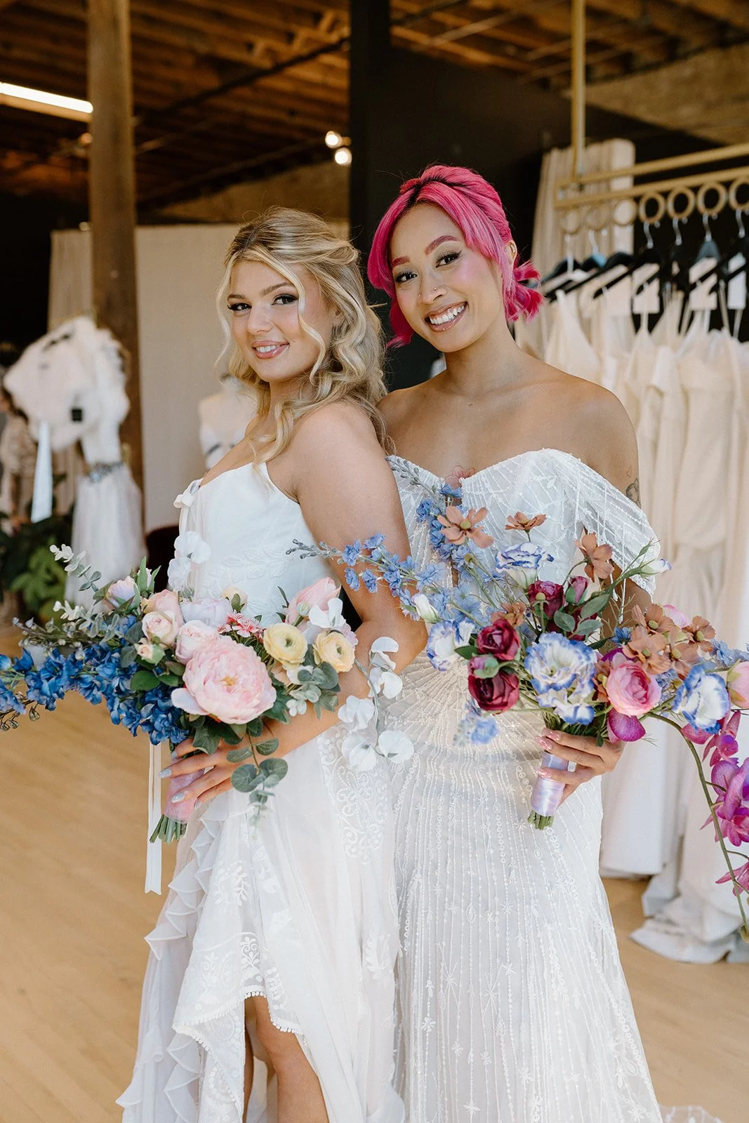 Two women in white dresses holding colorful bouquets of flowers, smiling, in what appears to be a bridal boutique or dress shop.