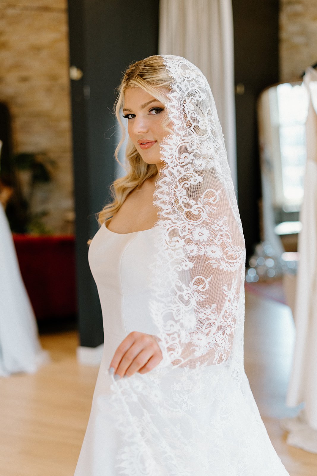 A bride in a white wedding dress with a lace veil, standing indoors with a blurred background of curtains and wooden walls.