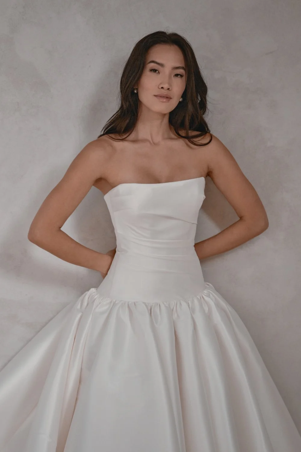 Woman wearing a strapless white satin ball gown, with a fitted bodice and full skirt, posing against a plain wall.
