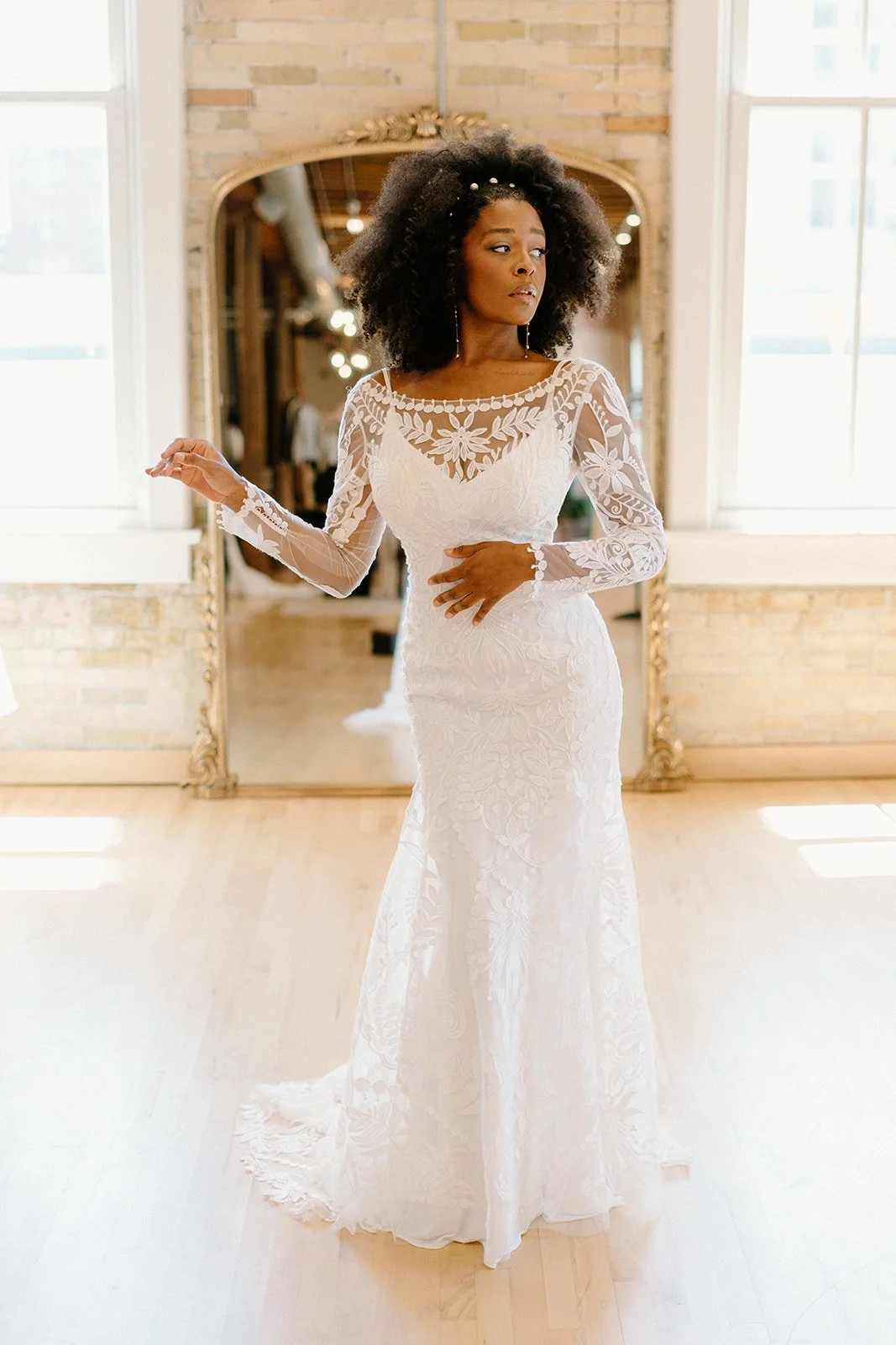 A woman in a white lace wedding dress with long sheer lace sleeves, standing in a room with large windows, a brick wall, and a large mirror behind her.