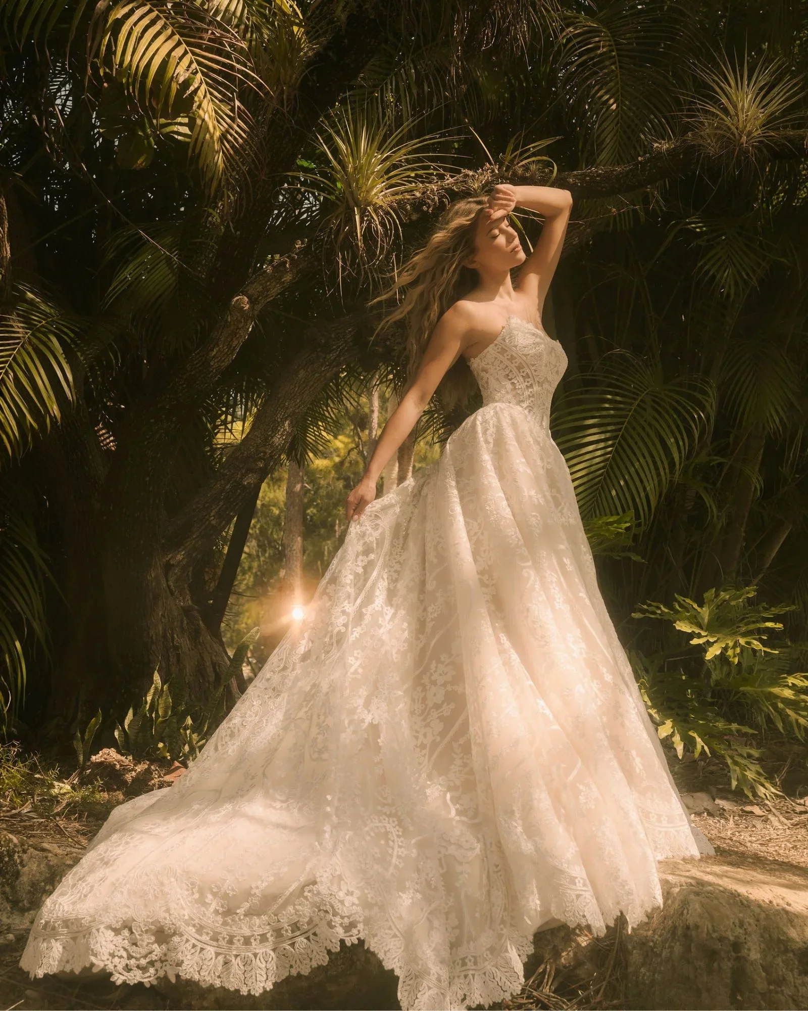 A woman in a white lace wedding gown standing outdoors among green tropical plants, with her eyes closed and one hand raised to her forehead, illuminated by warm sunlight.
