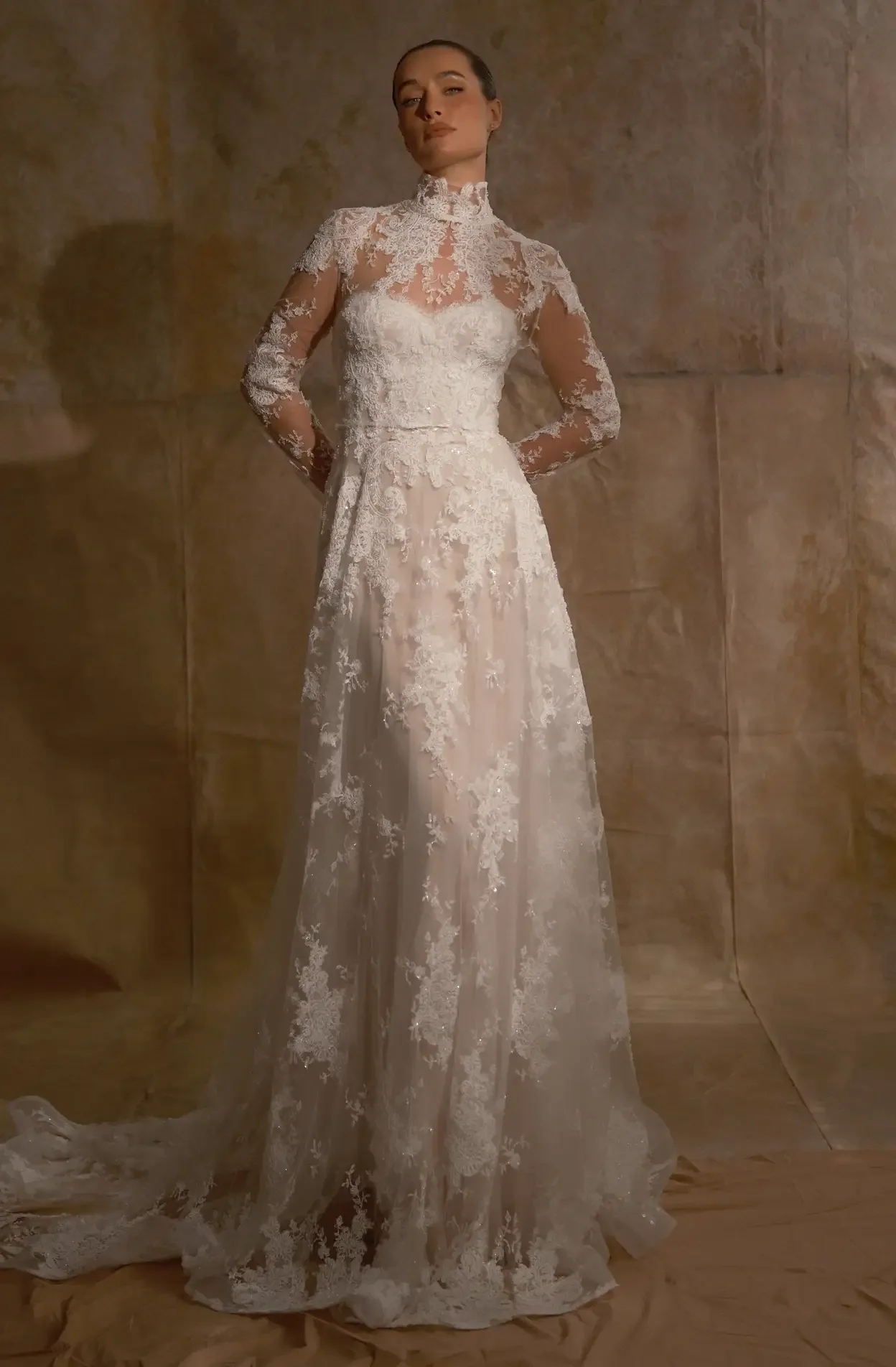 A woman in a long, white lace wedding dress standing against a stone wall.